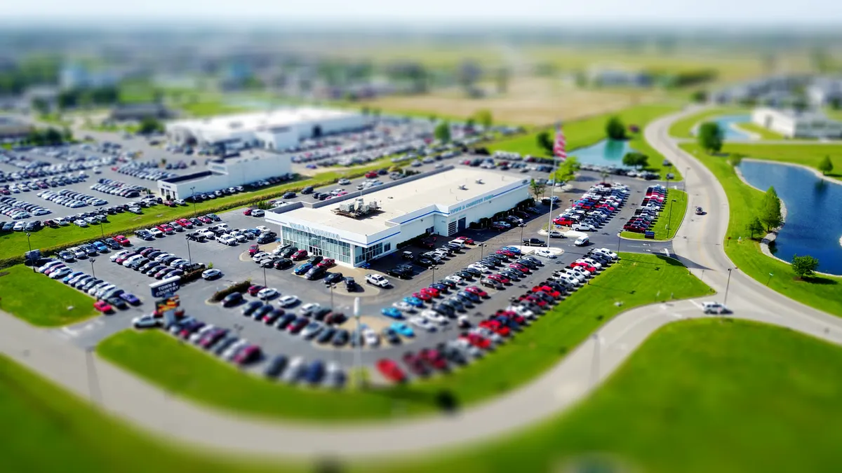 Aerial high-angle view of a bustling car dealership surrounded by parked cars in a green landscape.