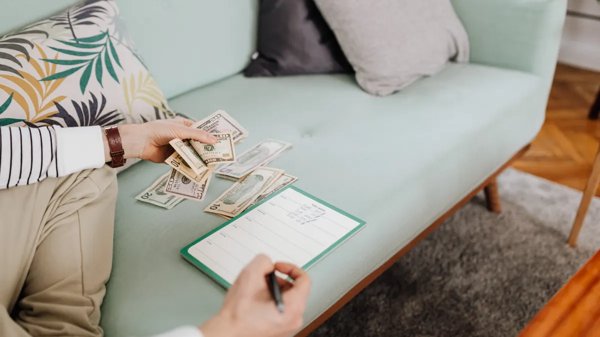 Adult counting cash and writing in a notebook while seated on a stylish couch at home.